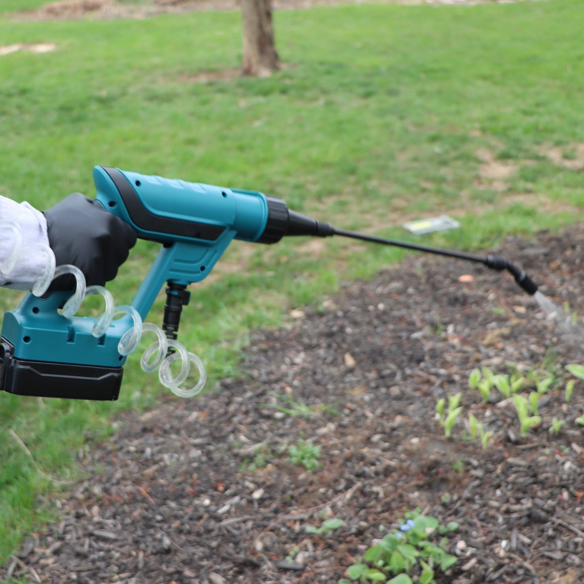 gloved hand holding battery powered spray gun spraying a garden bed